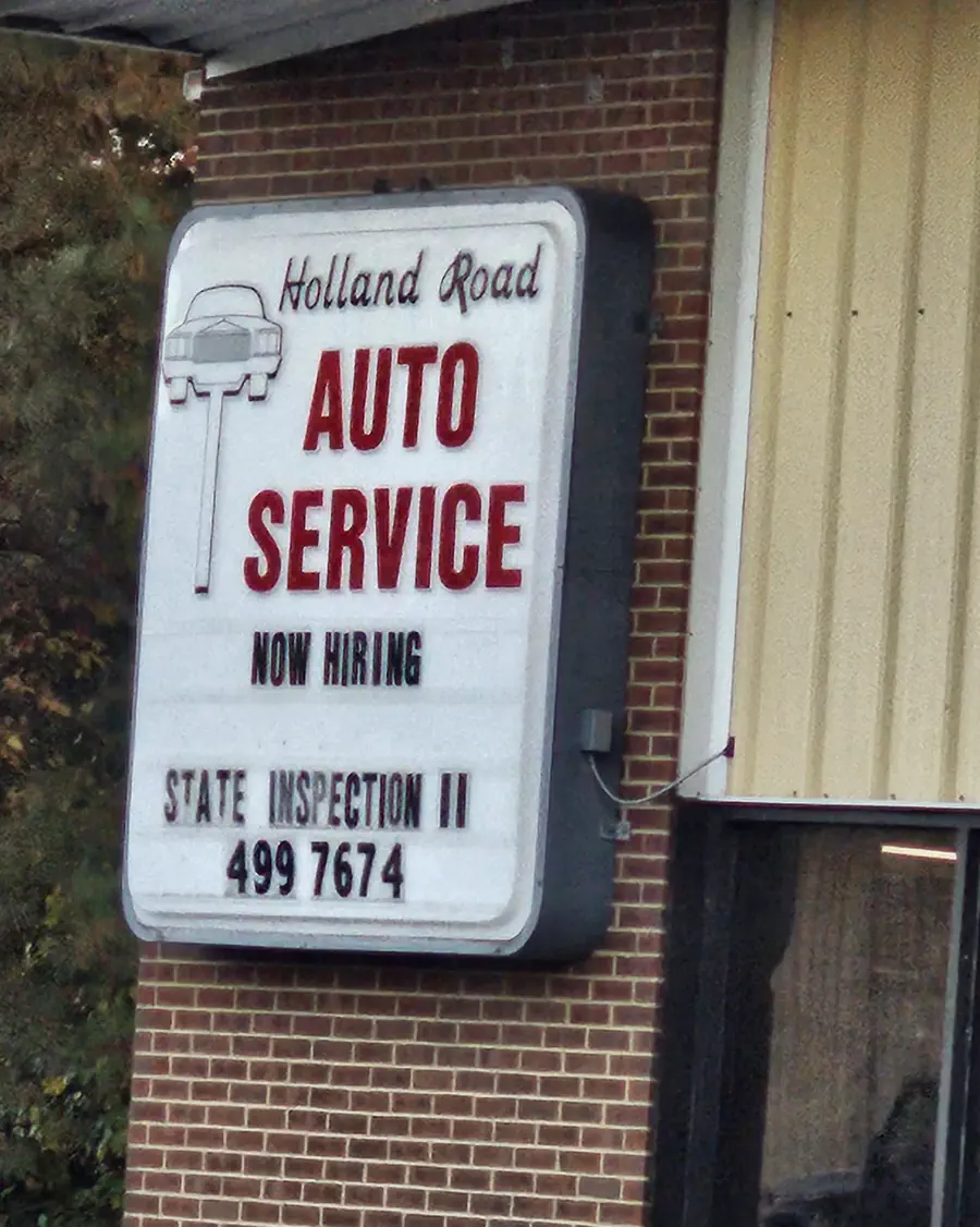 The lit Holland Road Auto Service street sign on the brick office, reading State Inspection and 499 7674